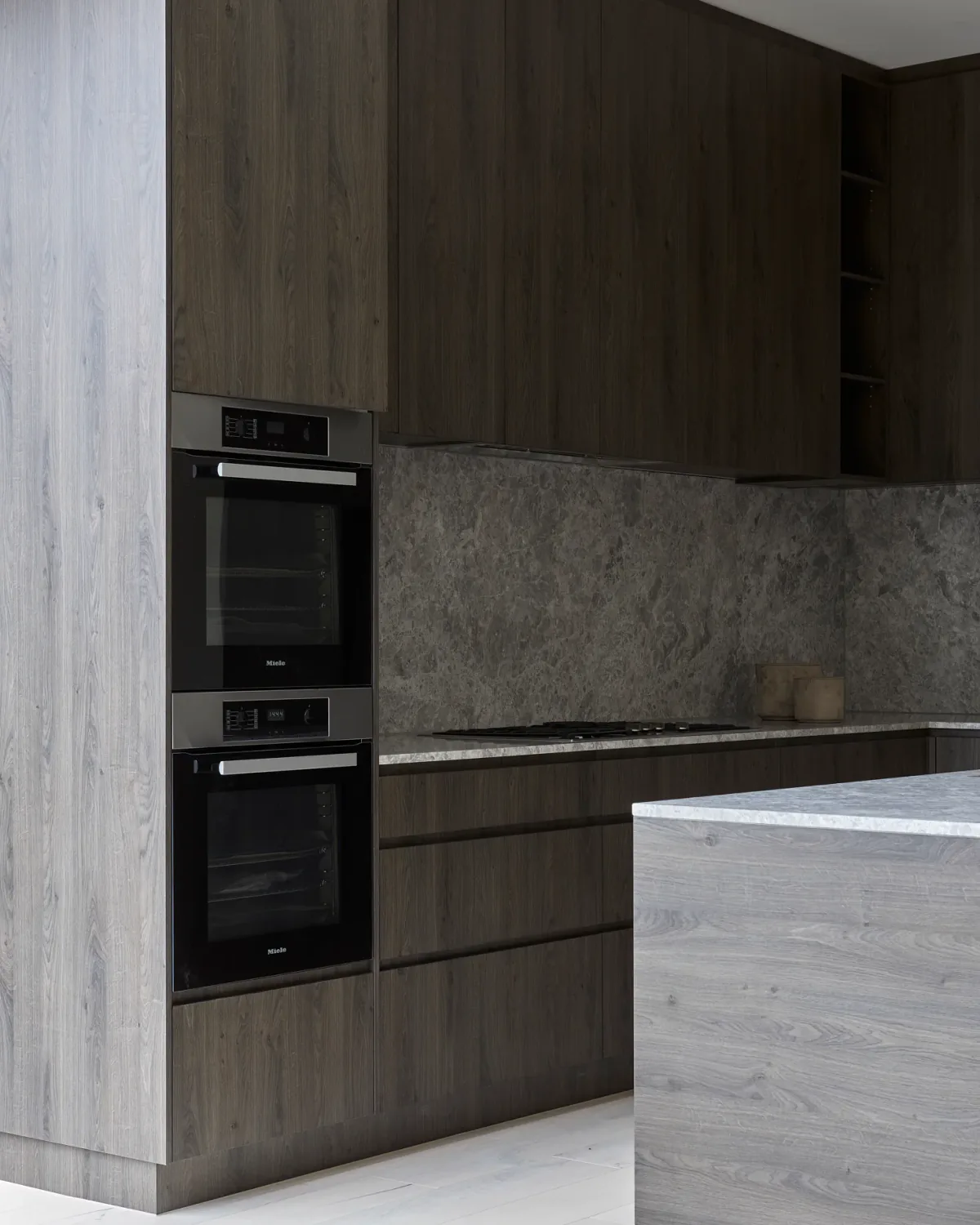 Dark kitchen cabinetry with integrated ovens, stone backsplash, and a light wood island.