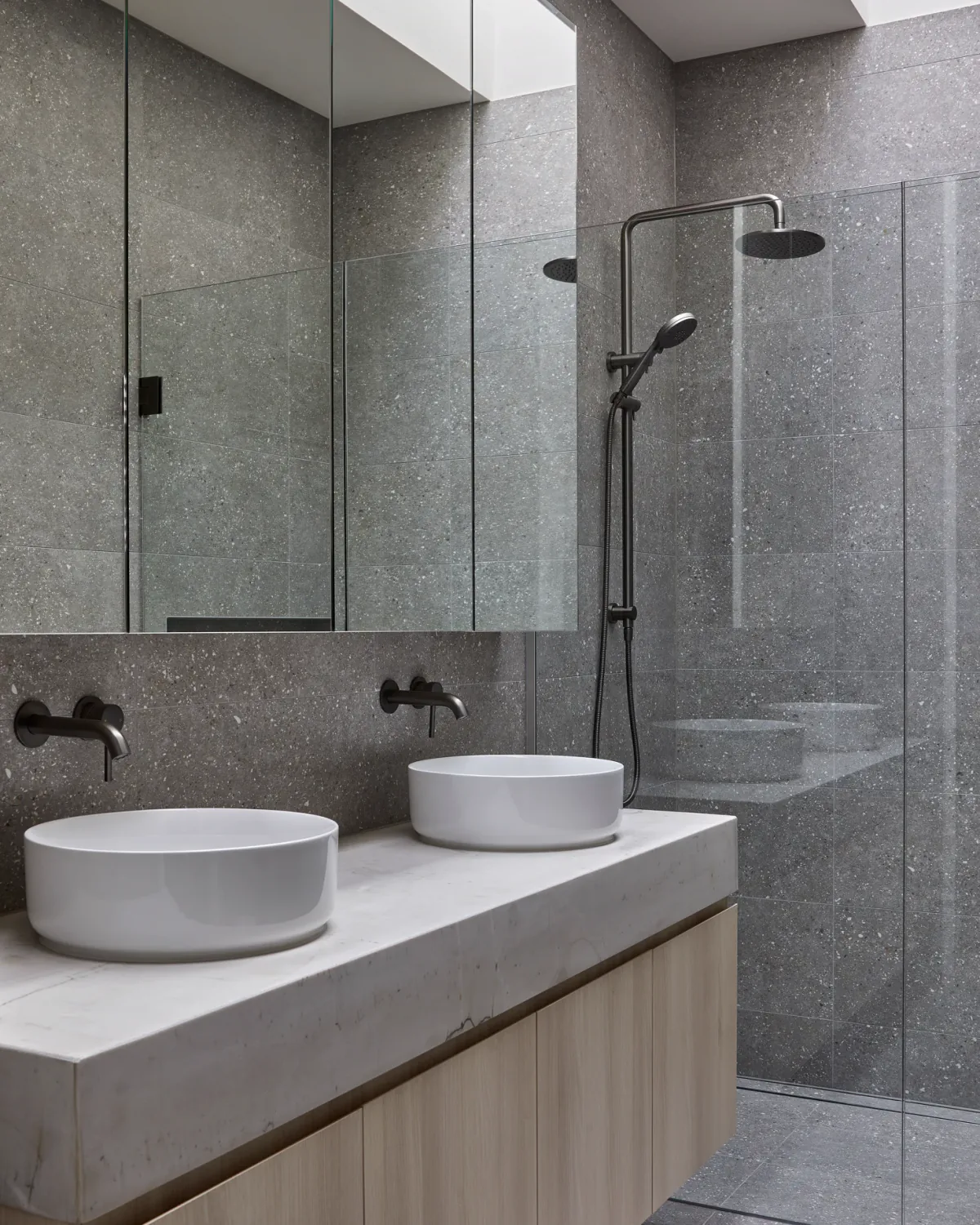 Gray tile bathroom with double vessel sinks, mirror cabinets, and a glass shower.