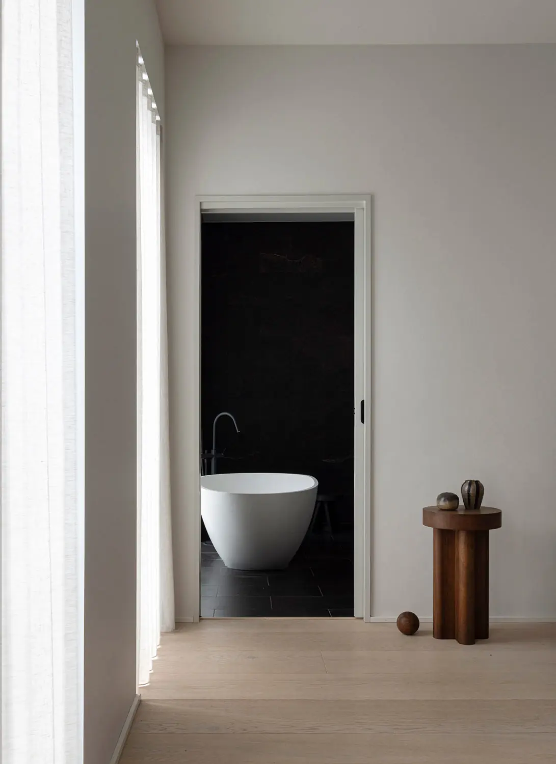 Bathroom view through a quiet doorway with freestanding white tub, dark tile, sheer curtains, and warm wood side table